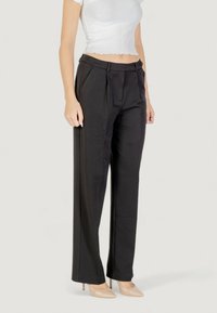 Pantaloni neri su misura con pieghe frontali, gamba larga e una texture liscia. Abbinati a tacchi a spillo beige su uno sfondo neutro.