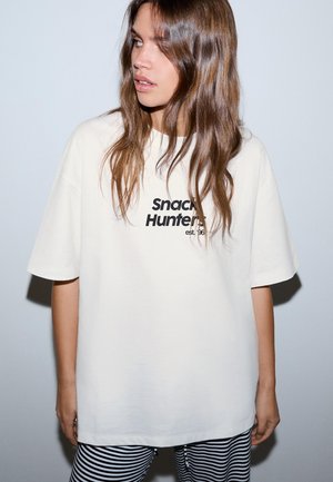 Mlada ženska z dolgimi rjavimi lasmi nosi preveliko belo majico s napisom "Snack Hunters est. '96" in črno-bele črtaste hlače.