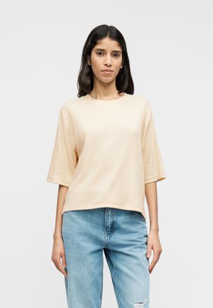 Camiseta de gran tamaño color beige claro con mangas cortas, confeccionada con tela suave, con cuello redondo y un corte holgado. Combinada con vaqueros azules.
