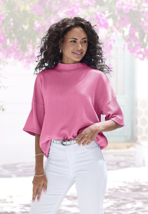 Rosa strukturiertes Oberteil mit hohem Kragen und kurzen Ärmeln, kombiniert mit weißen Jeans und einem hellen Gürtel. Der Hintergrund zeigt rosa Blumen.