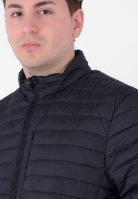 Zwarte gewatteerde jas met een staande kraag en horizontale quilt-ontwerp. Het materiaal lijkt glad met een matte afwerking.