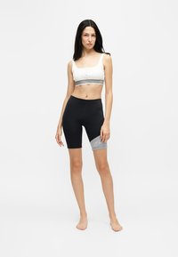 Femme debout pieds nus portant un crop top blanc et un short de cyclisme noir avec une bande grise avec logo sur la cuisse, sur fond uni.
