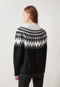 Svart genser med hvite geometriske mønstre, ribbestrikket krage og kant, samt en myk tekstur. Designet med en avslappet passform og lange ermer.