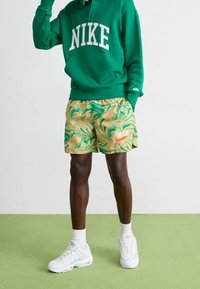 Grön huvtröja med vitt Nike-logotyp, mångfärgade marmorerade shorts i guld och grönt med en liten Nike-logotyp, vita sneakers och knästrumpor.