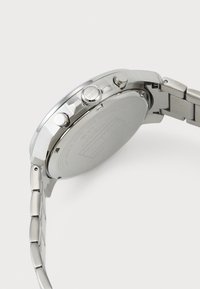 Silverfärgad klocka i rostfritt stål med rund boett, texturerad krona och två tryckknappar; länkad metallarmband som visar en polerad och borstad yta.