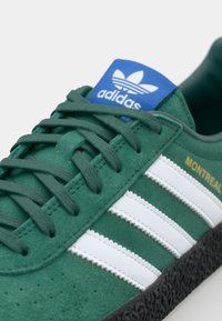 Adidași verzi din suede cu dungi albe, etichetă albastră pe limbă, talpă texturată neagră și detaliu auriu "MONTREAL". Au șireturi plate, împletite.