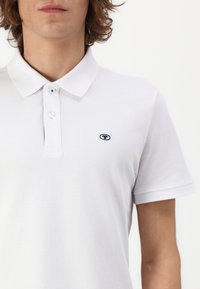 Tricou polo alb dintr-un material texturat, cu două butoane la guler și un mic logo bleumarin pe partea stângă a pieptului. Mâneci scurte.