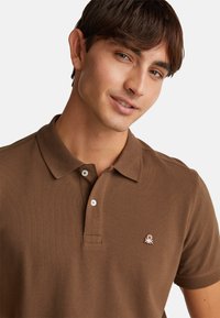 Hnědá polo košile vyrobená z texturované látky, s límečkem a dvouknoflíkovým zapínáním, malým vyšívaným logem na hrudi.