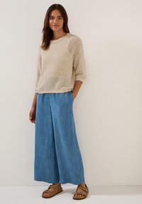 Beiger Strickpullover mit lockerer Passform, kombiniert mit hellblauen Jeans mit weitem Bein. Braune Sandalen mit verstellbaren Riemen runden das Outfit ab.