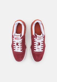 Adidași roșii din suede, cu șireturi albe și un interior portocaliu căptușit. Au o talpă intermediară albă și o talpă din cauciuc pentru aderență. Logo-ul Reebok este vizibil.
