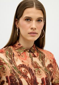 Chemise à manches longues et col, ornée d'un motif abstrait coloré dans des tons de pêche, vert et marron, avec une fermeture à boutons au niveau du cou.