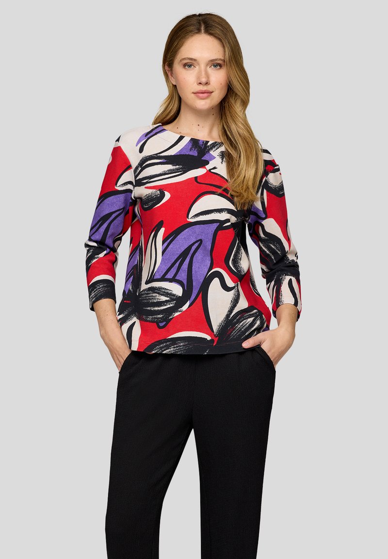 Floral-patterned top with red, purple, and black design on a light background. Three-quarter sleeves and round neckline. Paired with black pants.