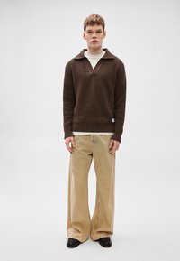 Barna kötött pulóver széles gallérral, krémszínű ing fölött viselve. Széles szárú bézs nadrággal és fekete cipővel párosítva.