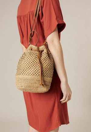 Personne portant une robe couleur rouille, portant un sac seau tissé beige avec un cordon de serrage en cuir et des bretelles réglables sur le dos.