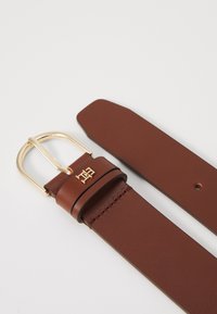 Brown leather belt with a gold metal buckle, featuring a branded detail and a smooth texture; tapers towards the end with a cut edge.