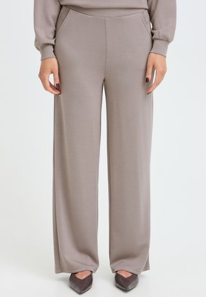 Femme portant un pantalon large taupe avec des poches latérales et un haut assorti, associé à des chaussures pointues marron foncé sur un fond uni.
