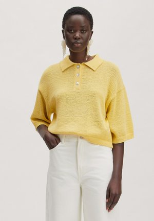 Vrouw draagt een los geel gebreid poloshirt met drie knopen en witte hoge taillebroeken, staand tegen een effen achtergrond.
