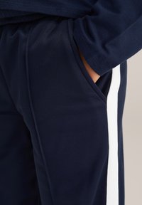 Roka iebāzta tumši zilos biksēs ar baltu sānu svītru, parādot auduma tekstūru un šuvju detaļas.