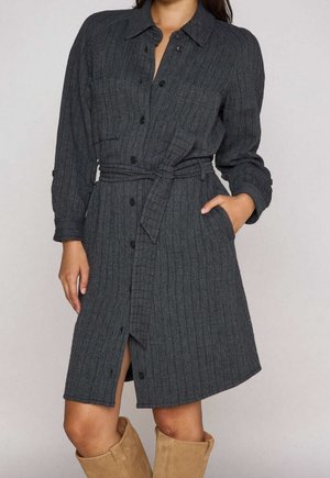 Donna che indossa un vestito grigio scuro, a coste, abbottonato, con cintura in vita e stivali marrone chiaro al ginocchio, in piedi con una mano in tasca.