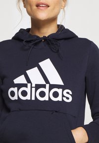 Granatowa bluza z kapturem wykonana z mieszanki bawełny, z białym logo Adidas, regulowanym sznurkiem i przednią kieszenią kangurką.