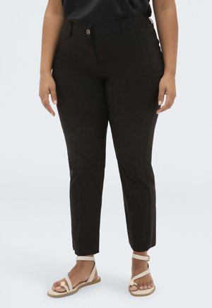 Femme portant un pantalon noir ajusté longueur cheville et des sandales beiges à bout ouvert, debout devant un fond clair uni.