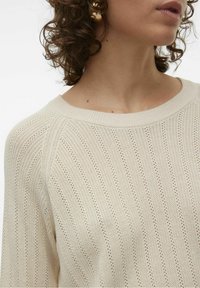 Personne aux cheveux bouclés portant un pull léger en tricot couleur crème avec un large col rond.