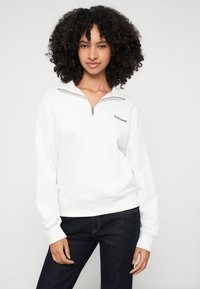 Donna con capelli ricci che indossa una felpa bianca con zip a quarto e il logo "GUESS JEANS" e jeans blu scuro, in piedi contro uno sfondo chiaro e uniforme.