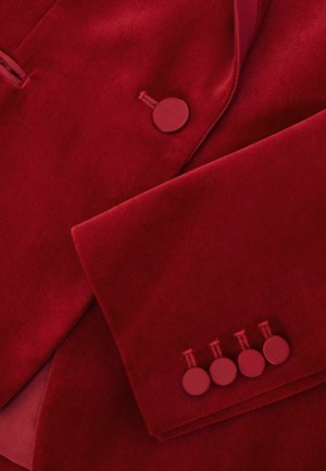 Piros bársonykabát sima textúrával, kerek gombokkal és szabott hajtókával. Négy azonos gomb díszíti az ujjak végét.