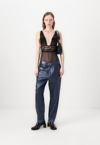 Bodysuit de encaje negro con escote en V profundo, combinado con pantalones sueltos de un azul oscuro brillante. La modelo lleva botas de tacón grueso negras y un bolso pequeño.