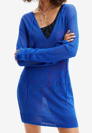 Femme portant une robe en maille bleue transparente à manches longues sur un haut noir, bras croisés, et un collier argenté avec un pendentif en forme de cœur.