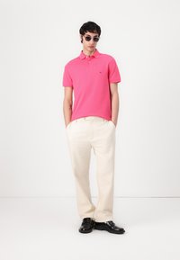 Rosa Poloshirt mit einem Kragen, kurzen Ärmeln und einem Logo auf der linken Brust. Kombiniert mit cremefarbenen Hosen und schwarzen eleganten Schuhen.