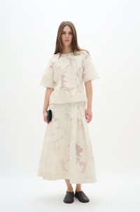 Durchsichtige cremefarbene Bluse und langer Rock mit floralem Muster, kombiniert mit schwarzen Slippern, vor einem schlichten weißen Hintergrund präsentiert.