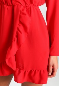Vestido rojo de tejido suave y fluido con un dobladillo con volantes y cinturón. El diseño incluye mangas largas y una cintura fruncida para aportar estructura.