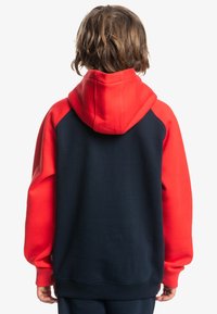 Hoodie rouge et bleu marine avec une poche kangourou, texture douce et poignets côtelés ; présente une capuche et un design en blocs de couleurs.