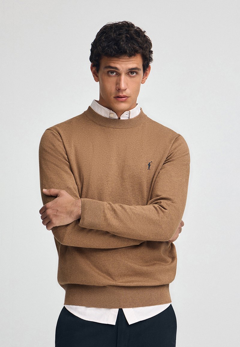 Braun gestrickter Pullover mit Rundhalsausschnitt und gerippten Bündchen, getragen über einem weißen Hemd mit feinen Streifen, das ein kleines Logo auf der Brust hat.