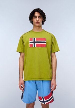 Giovane con capelli ricci che indossa una maglietta verde e pantaloncini blu, entrambi con disegni della bandiera rossa, bianca e blu.