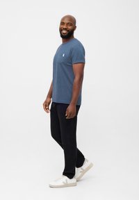 Blauwe katoenen t-shirt met een borstzak, gecombineerd met zwarte broeken en witte sneakers met lichte accenten. Model staat voor een neutrale achtergrond.