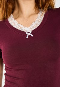 Burgundské fitted top s hlubokým V-výstřihem. Bílá krajka na límci má malou mašli. Hladká, elastická textilie.