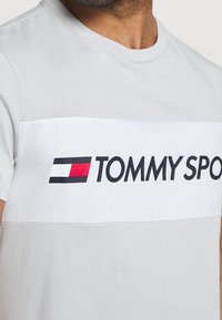Camiseta de algodón gris con una banda horizontal blanca que presenta un logo "TOMMY SPORT" en negro y rojo. Diseño de mangas cortas y cuello redondo.