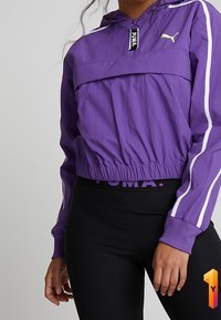 Purple cropped windbreaker with a half-zip, PUMA logo, white stripes on sleeves, and elastic waistband, paired with black ribbed leggings.