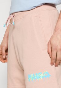 Shorts en coton rose en tissu doux, avec une taille élastique à cordon de serrage aux embouts bleus et un logo graphique multicolore sur le côté.