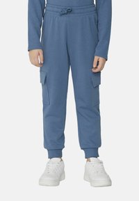 Modré bavlněné tepláky s cargo kapsami, elastickým pasem a ribbovanými manžetami. V kombinaci s bílými teniskami na neutrálním pozadí.