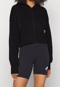 Svart kort zip-up hoodie med huva, i en slät tygkvalitet och diskreta sömnadsdetaljer, tillsammans med figurnära svarta shorts med logotypdetaljer.