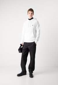 Sudadera blanca con capucha y un logo negro, combinada con pantalones de mezclilla sueltos de color gris oscuro y botas negras, sosteniendo una bolsa negra.