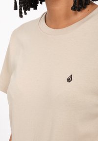 T-shirt beige a maniche corte realizzato in cotone. Presenta un piccolo emblema nero a forma di foglia sul lato sinistro del petto. Tessuto morbido e scollo rotondo.