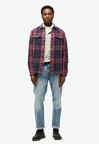 Geruit overhemd in rood, marineblauw en wit, gedragen over een grijze sweatshirt. Gecombineerd met lichtblauwe jeans en zwarte schoenen. Heeft borstzakken.