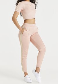 Světle růžový crop top a ladící tepláky se zlatými bočními pruhy. Materiál se zdá být měkký a elastický, s bočními kapsami. Bílé tenisky dokončují vzhled.