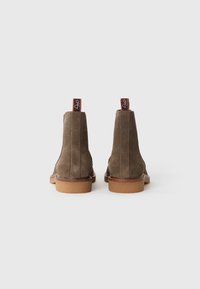 Un par de botas de tobillo de ante marrón con suelas de crepé en color marrón claro, vistas desde atrás sobre un fondo blanco.