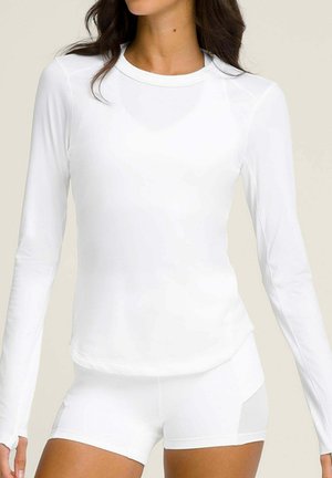 Donna che indossa una maglia bianca aderente a maniche lunghe e pantaloncini bianchi attillati abbinati, su uno sfondo neutro.