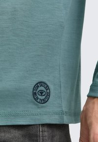Camisa de manga larga azul verdosa con dobladillo y parche circular de logo de Tom Tailor, combinada con jeans gris oscuro, y una mano masculina visible en el lado derecho.